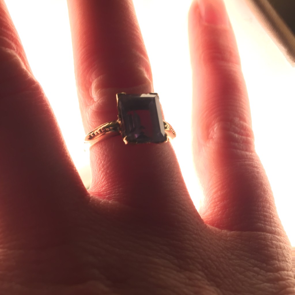 A woman's right hand wearing a gold ringt with an amethyst gemstone.